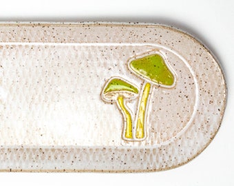 May include: An oval ceramic serving tray with a speckled, off-white surface. Two stylized mushrooms with bright green caps and yellow stems are featured on the right. The tray is a decorative item.