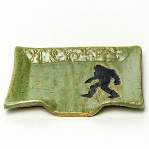 May include: A handmade, rectangular, green ceramic dish with a black Bigfoot silhouette. The dish has a slightly curved shape and a textured surface. The top edge features a decorative floral pattern. The dish is likely used for serving or display.