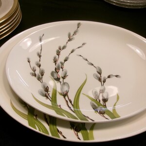 May include: Two white oval serving platters with a floral design. The design features gray pussy willows and green leaves. The platters are stacked on top of each other. Other white and gold rimmed plates are visible in the background.