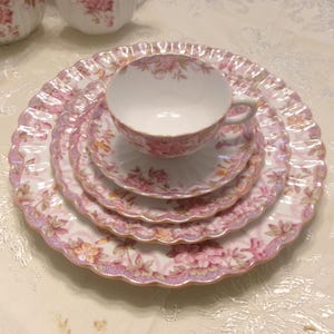May include: A porcelain tea set featuring a teacup, saucer, and two stacked plates with a scalloped edge. The set is decorated with a pink and white floral pattern, accented by a subtle iridescent finish along the wavy, gold-trimmed borders.