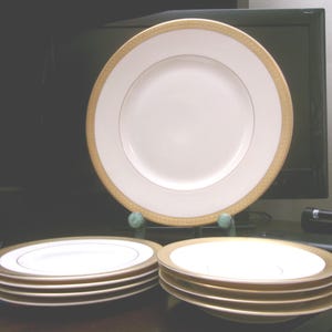 May include: A set of white porcelain plates with a gold border and a gold inner rim. The plates are stacked in two piles, with one plate displayed on a stand. The plates have a classic, elegant design.