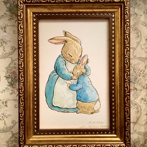 May include: A framed watercolor illustration of two rabbits, one larger and one smaller, in a tender embrace. The larger rabbit wears a blue dress, and the smaller rabbit wears a blue jacket. The frame is ornate gold.