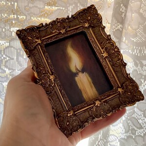 Puede incluir: Un peque&ntilde;o marco dorado adornado con una pintura de una vela encendida. La vela es blanca con una llama amarilla.