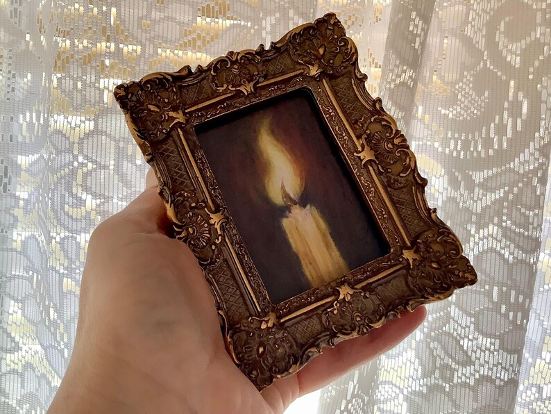 Puede incluir: Un peque&ntilde;o marco dorado adornado con una pintura de una vela encendida. La vela es blanca con una llama amarilla.
