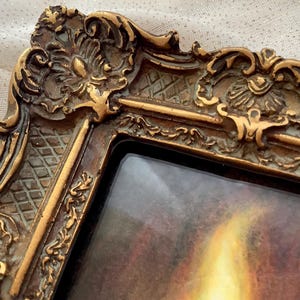 Puede incluir: Un marco de imagen dorado adornado con un dise&ntilde;o detallado. El marco tiene un frente de vidrio y un passe-partout marr&oacute;n oscuro. El marco est&aacute; vac&iacute;o.