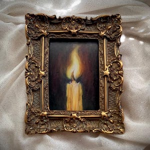 Puede incluir: Una pintura enmarcada de una vela encendida en un entorno oscuro. La vela es blanca y la llama es amarilla. El marco es ornamentado y dorado.
