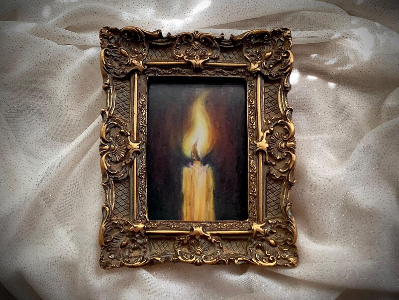 Puede incluir: Una pintura enmarcada de una vela encendida en un entorno oscuro. La vela es blanca y la llama es amarilla. El marco es ornamentado y dorado.
