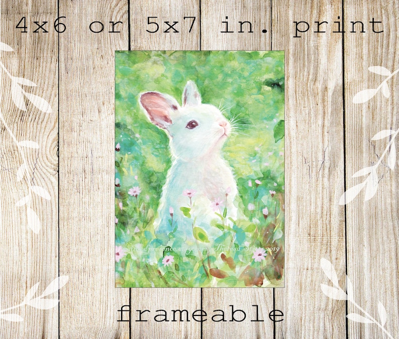 May include: A watercolor print of a white rabbit in a field of green grass and small pink flowers. The print is available in 4x6 or 5x7 inch sizes and is labeled "frameable". The rabbit is looking upwards with its ears perked.