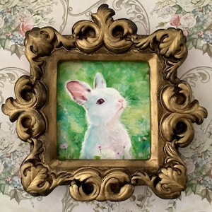 Può includere: Dipinto incorniciato di un coniglio bianco con orecchie rosa, su sfondo verde con fiori. La cornice dorata e ornata aggiunge un tocco vintage. L'opera evoca un tema primaverile e fantasioso, con un'atmosfera campestre.