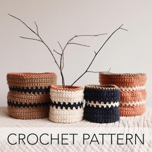 Puede incluir: Cuatro cestas de crochet en diferentes colores y patrones. Las cestas están hechas con hilo marrón, crema, negro y naranja. Las cestas están sobre una superficie blanca con una rama en el centro.