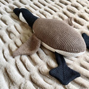 May include: A knitted plush goose toy in shades of brown, black, and white, lying on a textured beige surface. The toy features a detailed design with wings, a beak, and webbed feet. The goose is a soft, cuddly item.