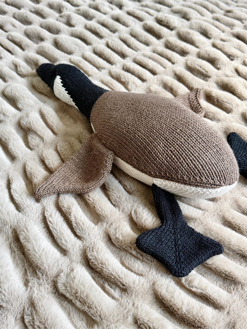 May include: A knitted plush goose toy in shades of brown, black, and white, lying on a textured beige surface. The toy features a detailed design with wings, a beak, and webbed feet. The goose is a soft, cuddly item.