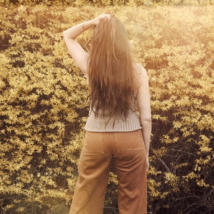 Peut inclure: Une femme avec de longs cheveux bruns se tient devant un mur de feuillage jaune. Elle porte un haut sans manches marron clair et un pantalon marron.