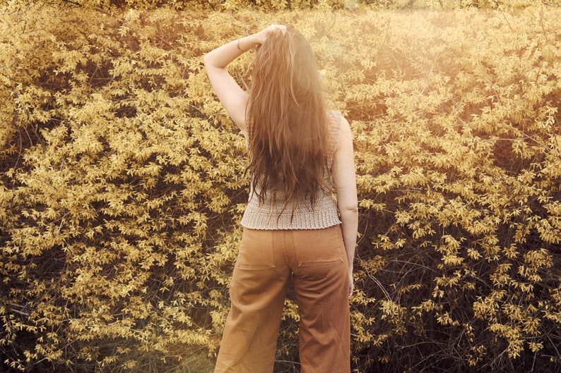 Peut inclure: Une femme avec de longs cheveux bruns se tient devant un mur de feuillage jaune. Elle porte un haut sans manches marron clair et un pantalon marron.
