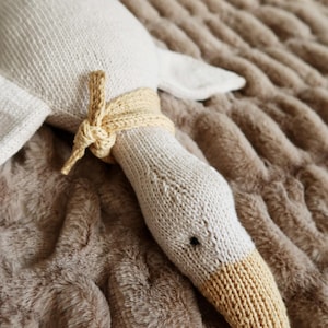 May include: A knitted goose plush toy in cream and tan. The goose has a long neck with a tan beak and a cream-colored body. A tan knitted band is tied around the neck. The toy is lying on a brown, textured surface.