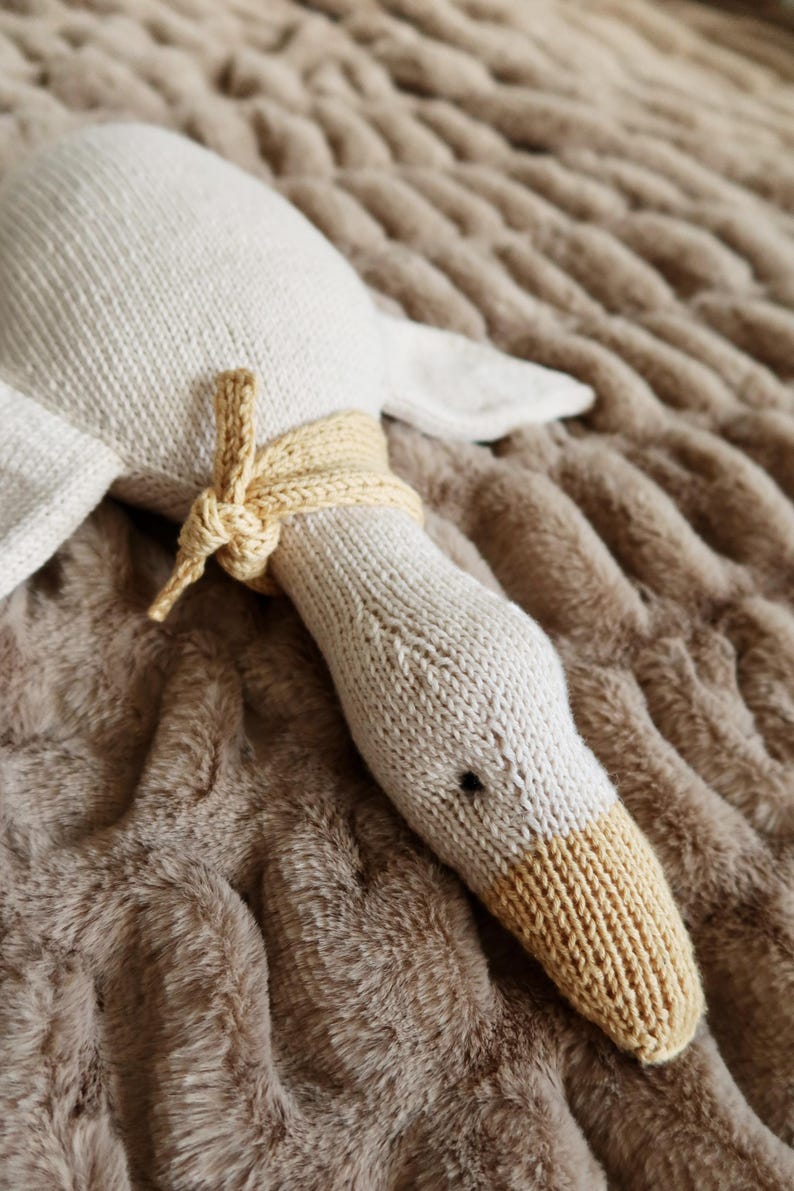 May include: A knitted goose plush toy in cream and tan. The goose has a long neck with a tan beak and a cream-colored body. A tan knitted band is tied around the neck. The toy is lying on a brown, textured surface.