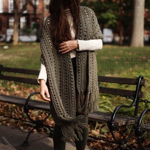 May include: A woman wearing a green crocheted shawl with fringe. The shawl is open in the front and has a loose, open weave. The woman is standing in front of a park bench.