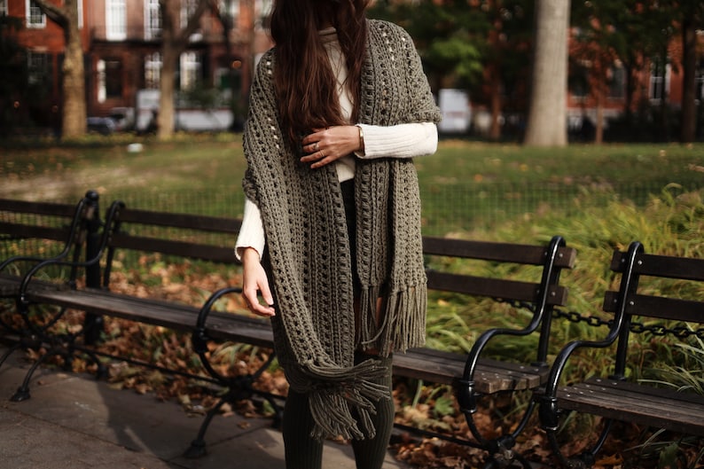 May include: A woman wearing a green crocheted shawl with fringe. The shawl is open in the front and has a loose, open weave. The woman is standing in front of a park bench.