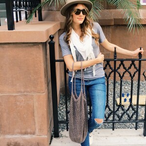 May include: A woman wearing a grey t-shirt, blue jeans, a brown hat, and sunglasses. She is carrying a brown crocheted tote bag and a white scarf.