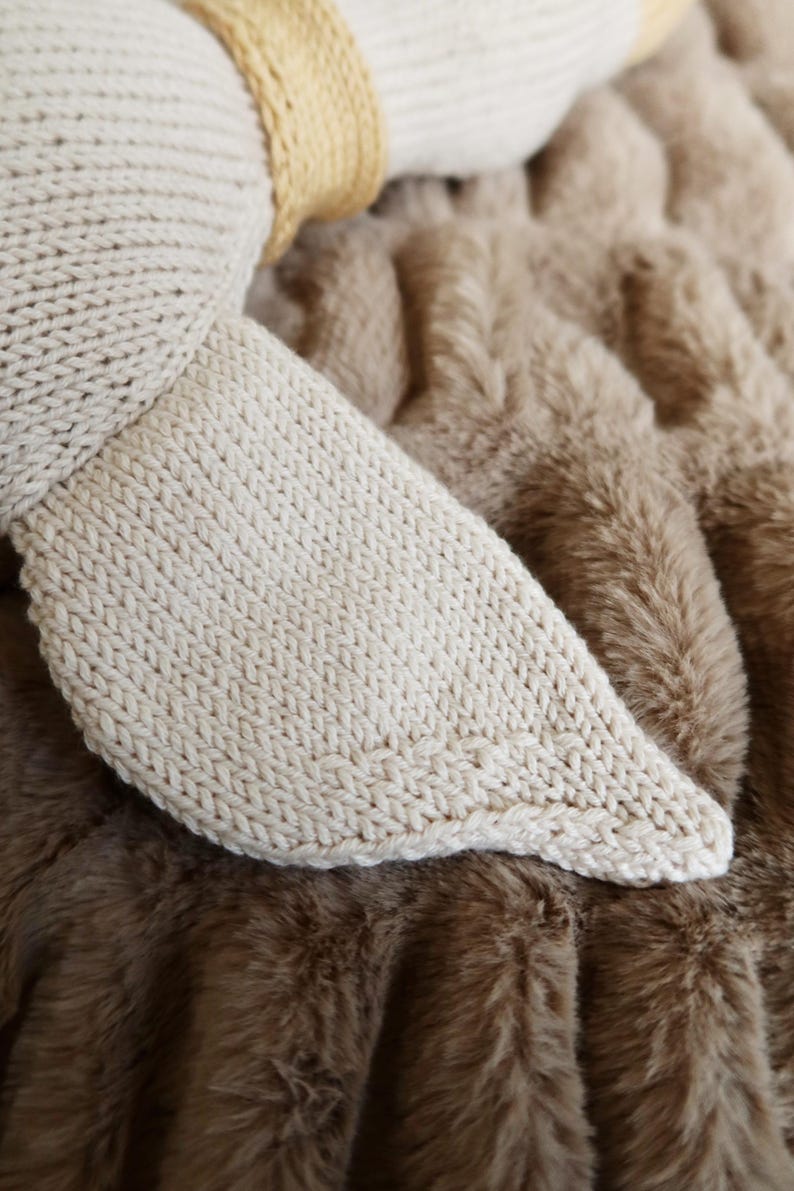 May include: Close-up of a knitted, off-white fabric, possibly a stuffed animal's ear or foot, resting on a brown, faux fur blanket. The knitted piece has a rounded shape and a textured surface.