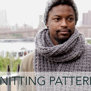 May include: A grey knitted scarf with a textured pattern. The scarf is worn by a person wearing a grey beanie and a brown jacket. The background is a blurry city skyline.