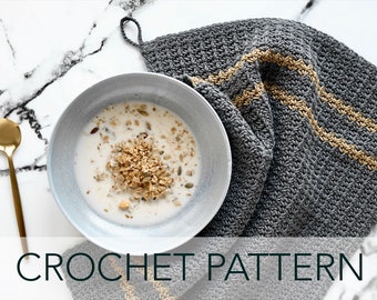 May include: A gray crochet dishcloth with two gold stripes. The dishcloth is folded and placed on a white marble surface. A bowl of cereal is in the background.
