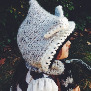 May include: A gray and white knitted hooded hat with a white fur trim. The hat is worn by a child with long brown hair. The child is wearing a white lace trim shirt and a black and white striped scarf.