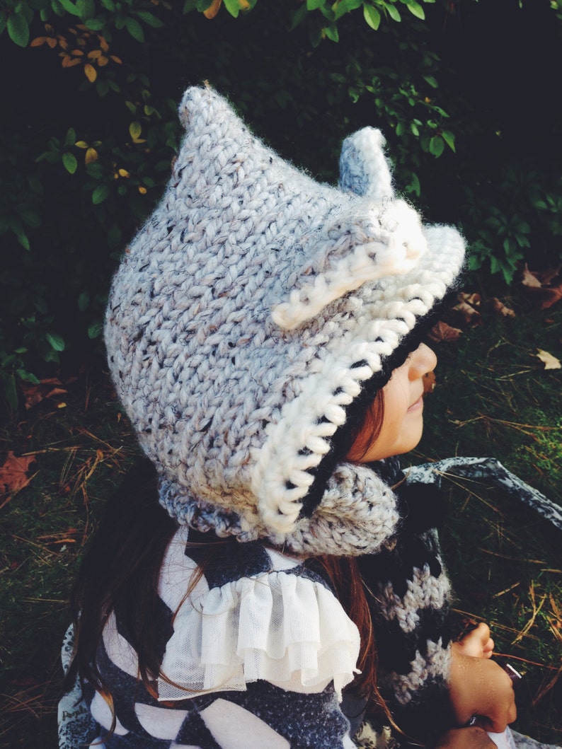 May include: A gray and white knitted hooded hat with a white fur trim. The hat is worn by a child with long brown hair. The child is wearing a white lace trim shirt and a black and white striped scarf.