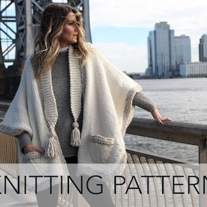 May include: A white knitted poncho with a gray trim and a matching scarf with a tassel. The poncho has pockets and a shawl collar. The model is wearing the poncho and scarf on a bridge with a city skyline in the background.