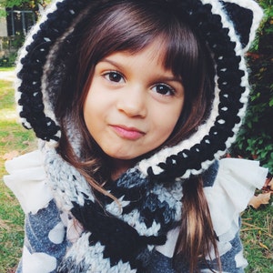 May include: A young girl wearing a black and white crocheted hooded scarf with a raccoon design. The scarf is worn with a gray and white patterned sweater.