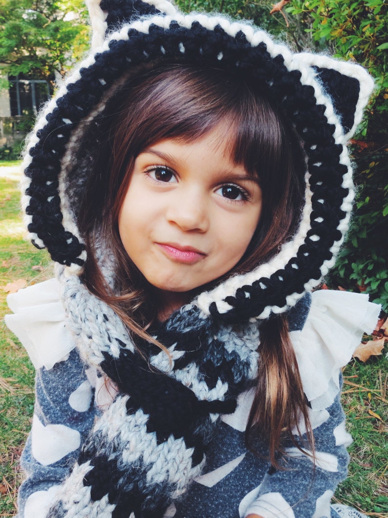 May include: A young girl wearing a black and white crocheted hooded scarf with a raccoon design. The scarf is worn with a gray and white patterned sweater.