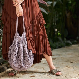 May include: A woman in a brown maxi dress holds a crocheted lavender tote bag. The bag has a large, open top and a wide, flat bottom.