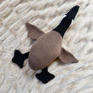 May include: A knitted goose toy in shades of brown, black, and white. The goose has a brown body, wings, and a black head with white markings. The feet are black. The toy is lying on a textured, light-colored surface.