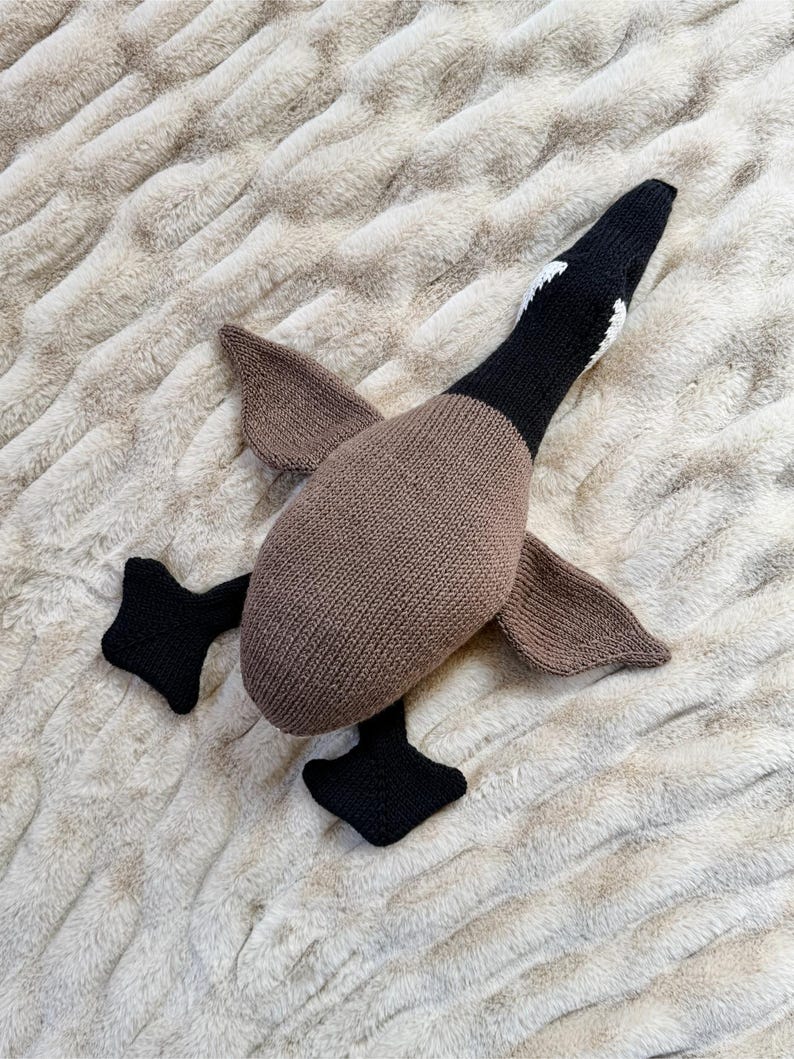 May include: A knitted goose toy in shades of brown, black, and white. The goose has a brown body, wings, and a black head with white markings. The feet are black. The toy is lying on a textured, light-colored surface.