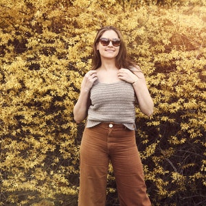 Peut inclure: Une femme portant un d&eacute;bardeur en crochet marron clair et un pantalon large marron. Elle est debout devant un buisson jaune et sourit.