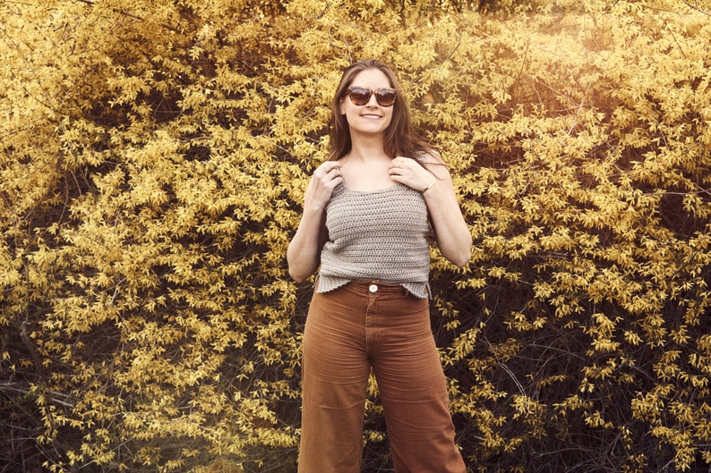 Peut inclure: Une femme portant un d&eacute;bardeur en crochet marron clair et un pantalon large marron. Elle est debout devant un buisson jaune et sourit.