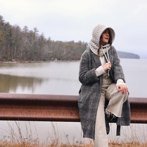 May include: A woman wearing a gray hooded sweater and a gray and black plaid coat sits on a rusty metal railing overlooking a lake. The lake is calm and the sky is overcast.