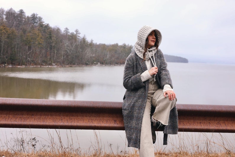 May include: A woman wearing a gray hooded sweater and a gray and black plaid coat sits on a rusty metal railing overlooking a lake. The lake is calm and the sky is overcast.