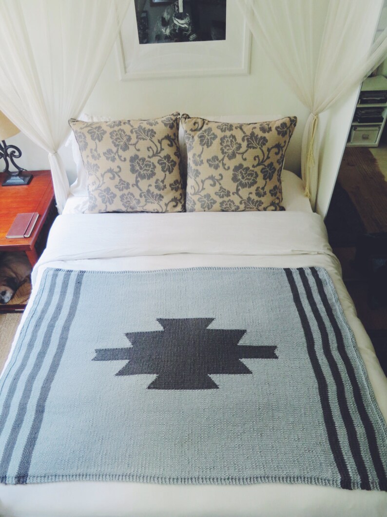 Puede incluir: Una manta de color azul claro con un patr&oacute;n geom&eacute;trico gris oscuro y rayas negras en los bordes. La manta est&aacute; sobre una cama blanca con dos almohadas estampadas.