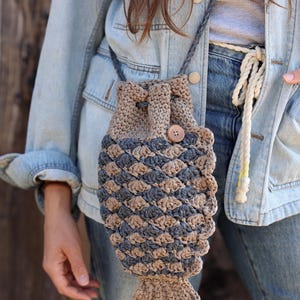May include: A handmade crochet bag in a fish shape, featuring a drawstring closure and a wooden button. The bag is primarily tan and blue, with a braided strap. The bag is worn over the shoulder.