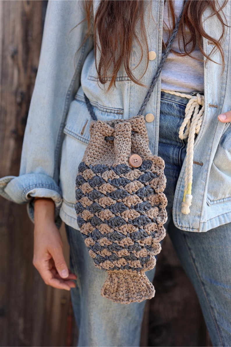 May include: A handmade crochet bag in a fish shape, featuring a drawstring closure and a wooden button. The bag is primarily tan and blue, with a braided strap. The bag is worn over the shoulder.