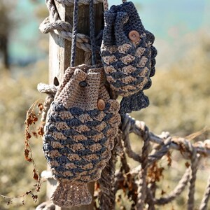 May include: Two handmade crochet fish-shaped bags in shades of tan and blue, hanging from a wooden post. The bags have a textured scale pattern and wooden button closures. A rope fence and blurred background complete the coastal theme.
