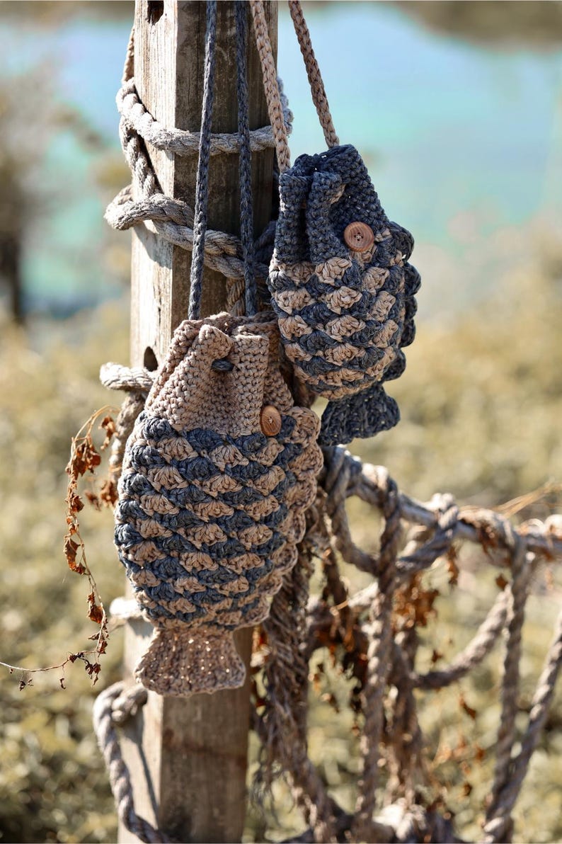 May include: Two handmade crochet fish-shaped bags in shades of tan and blue, hanging from a wooden post. The bags have a textured scale pattern and wooden button closures. A rope fence and blurred background complete the coastal theme.