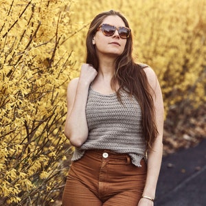 Peut inclure: Une femme portant un d&eacute;bardeur en crochet marron avec une ceinture marron et un pantalon marron. Elle se tient devant un buisson jaune et porte des lunettes de soleil.