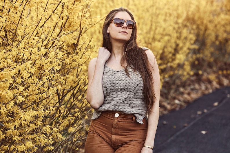 Peut inclure: Une femme portant un d&eacute;bardeur en crochet marron avec une ceinture marron et un pantalon marron. Elle se tient devant un buisson jaune et porte des lunettes de soleil.