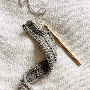 Peut inclure: Gros plan sur une bande de crochet gris avec un crochet en or et du fil gris. Le crochet est pos&eacute; sur une surface textur&eacute;e blanche.