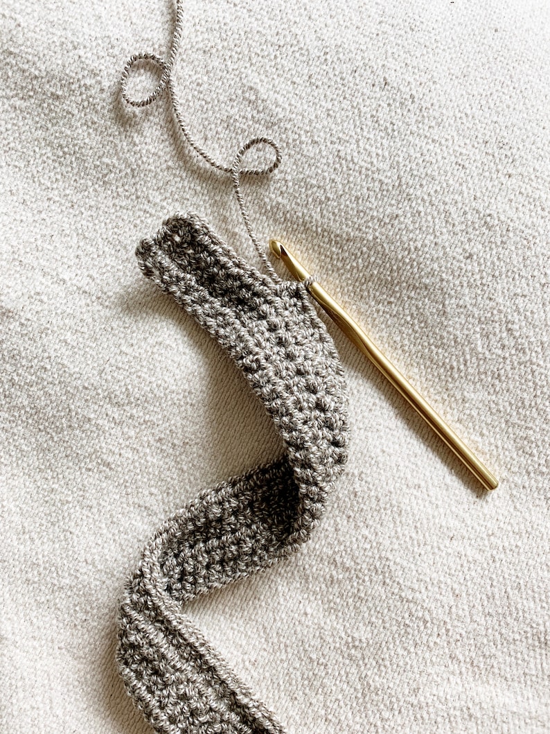 Peut inclure: Gros plan sur une bande de crochet gris avec un crochet en or et du fil gris. Le crochet est pos&eacute; sur une surface textur&eacute;e blanche.