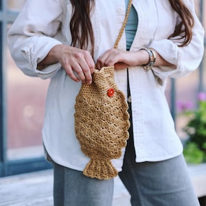 May include: A tan crochet fish-shaped crossbody bag with a drawstring closure and a red button. The bag has a textured, scale-like pattern and a long, woven strap. The bag is worn over the shoulder.