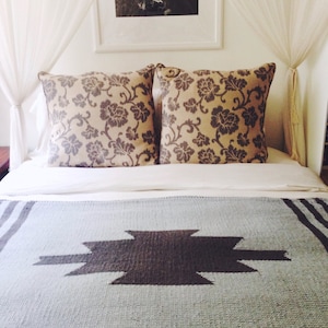 Puede incluir: Una cama blanca con una manta de patr&oacute;n gris y negro. La cama tiene dos almohadas con un patr&oacute;n floral. La cama est&aacute; en una habitaci&oacute;n con cortinas blancas.
