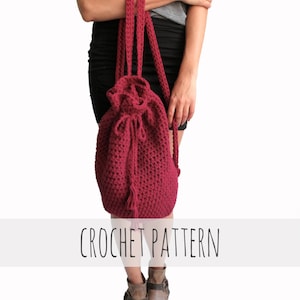 Könnte beinhalten: Ein maronfarbener gehäkelter Rucksack mit Kordelzug und langen Trägern. Die Tasche hat einen Quasten am Boden. CROCHET PATTERN steht in schwarzer Schrift auf weißem Hintergrund.